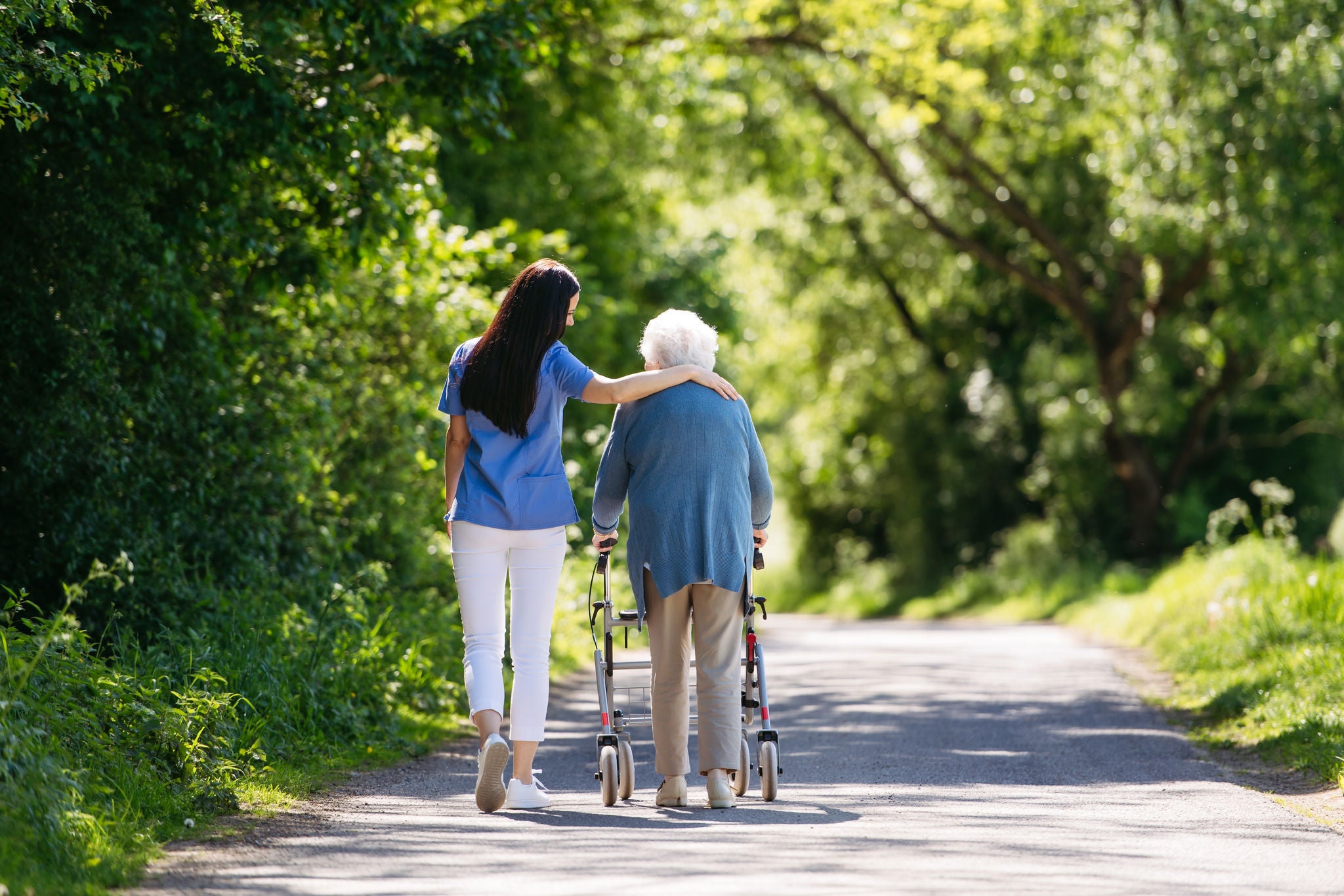Female caregiver and senior woman with walker on walk in nature. Nurse and elderly woman enjoying a warm day in nursing home, public park.