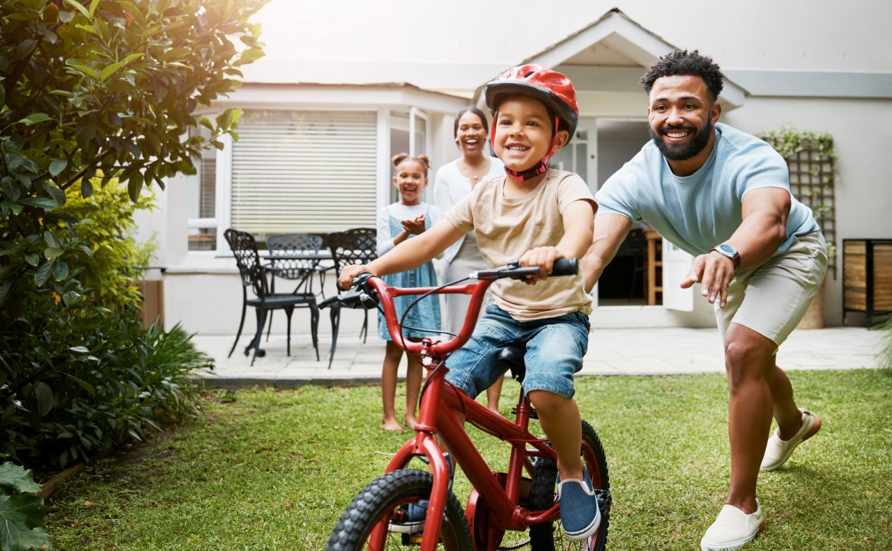 Learning, bicycle and proud dad teaching his young son to ride while wearing a helmet for safety in their family home garden. Active father helping and supporting his child while cycling outside